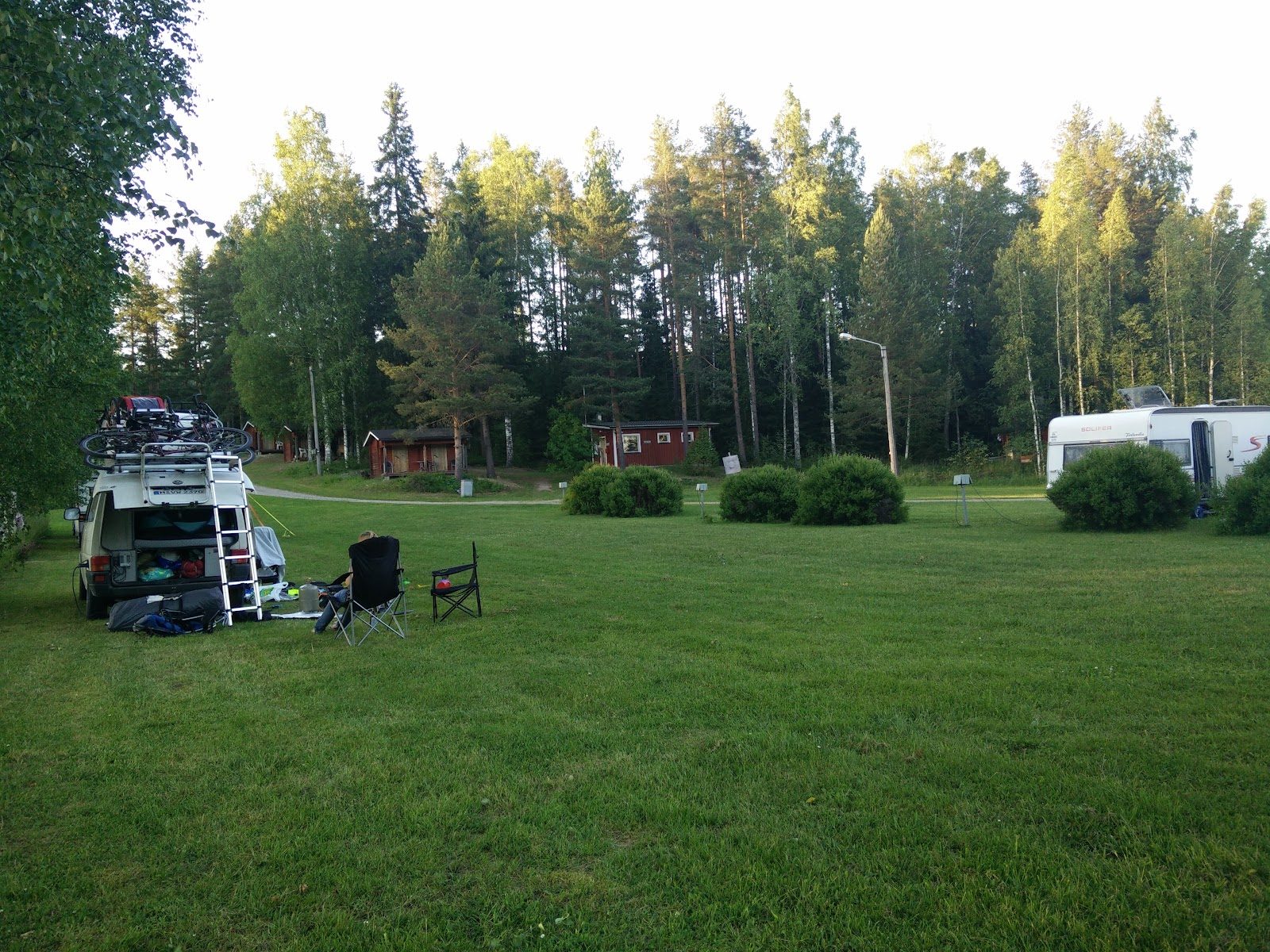 Nurmikenttä, jolla matkailuauto, retkituoleja ja telttoja. Taustalla puita ja vaunuja.