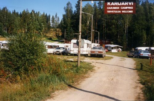 Vanha valokuva alueen sisäänajosta: sorapolku, pysäköityjä asuntovaunuja ja kyltti Karijärvi Caravan Camping, Tervetuloa.