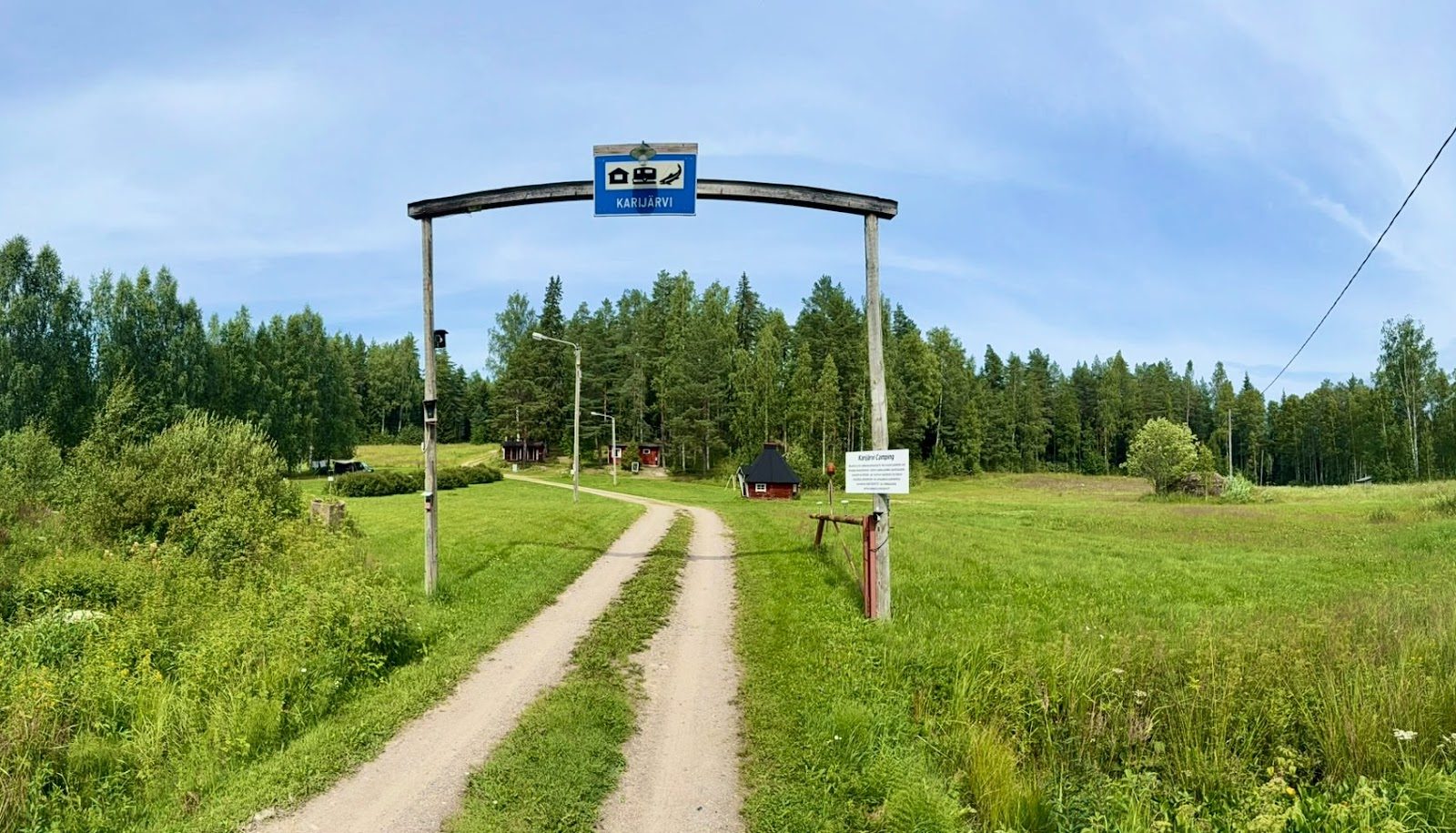 Karijärvi-kyltillä varustettu puuportti pellon laidalla, sorapolku vie kohti aittoja ja saunaa metsän reunassa.