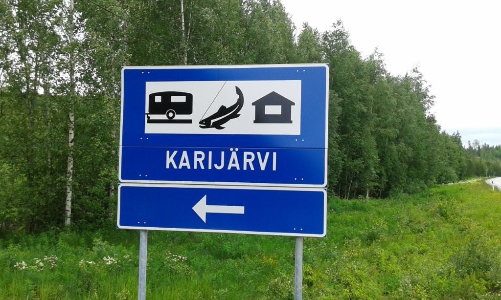 Sininen opastekyltti tien varressa: asuntovaunun, kalan ja mökin kuvat, teksti Karijärvi ja nuoli vasemmalle.