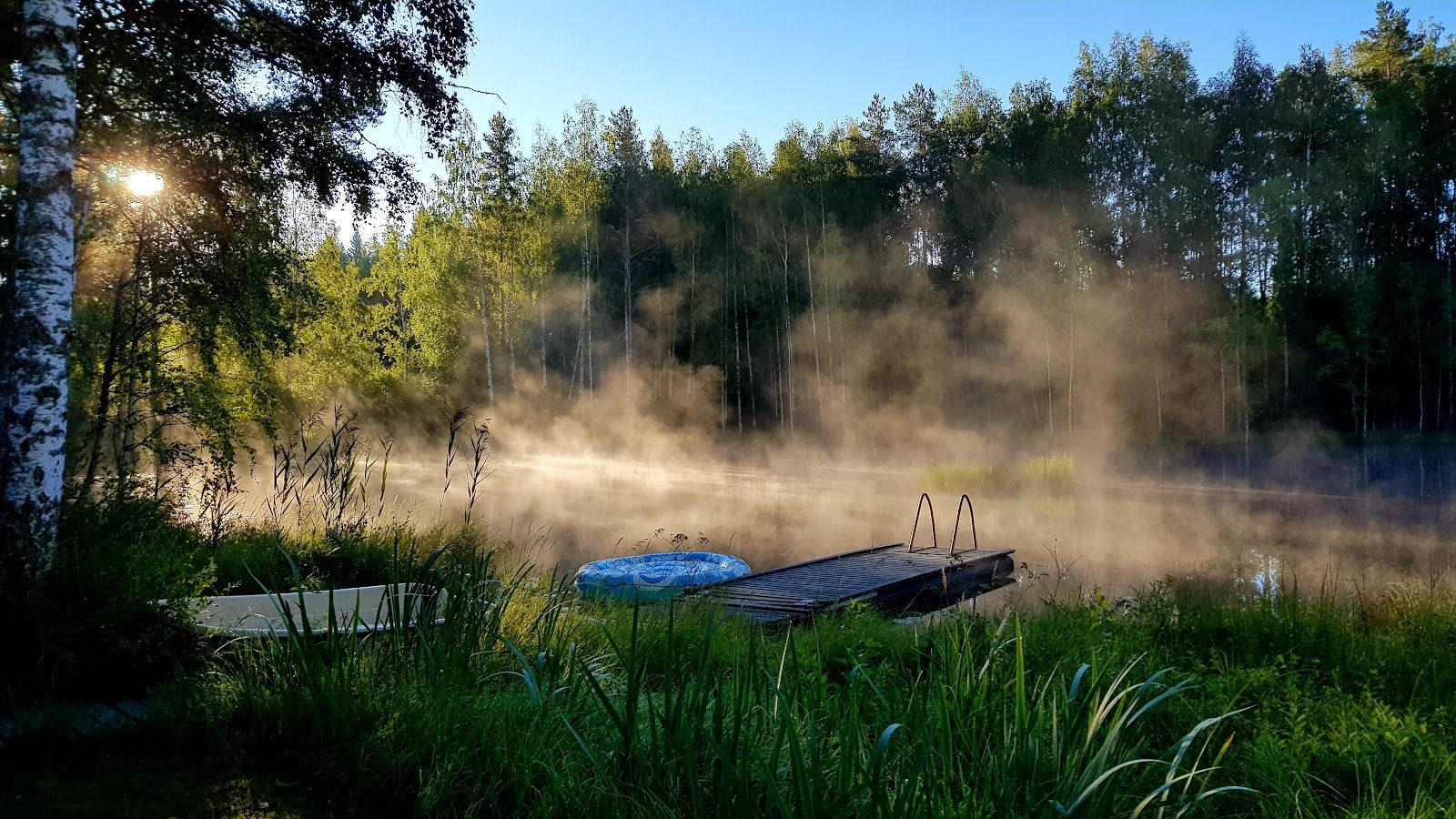 Usva nousee Karijärveltä aamuaurinkoon. Laituri ja vene rannassa, takana kuusikko.